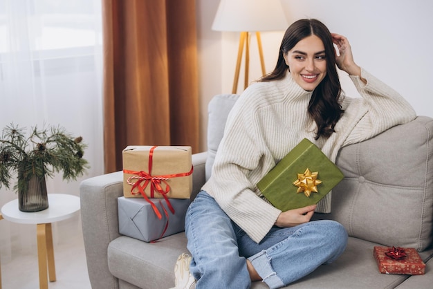 Beautiful happy woman unwrapping New Year or Christmas presents at home on sofa