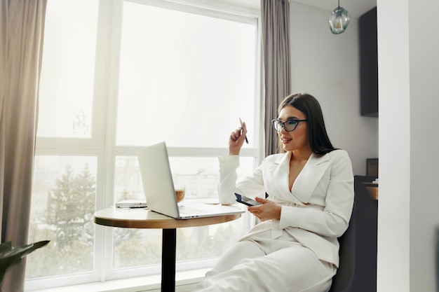 ノートパソコンで在宅勤務中に携帯電話を使用して美しい幸せな女性。スマートフォンでメッセージメッセージの眼鏡をかけている笑顔の女性。