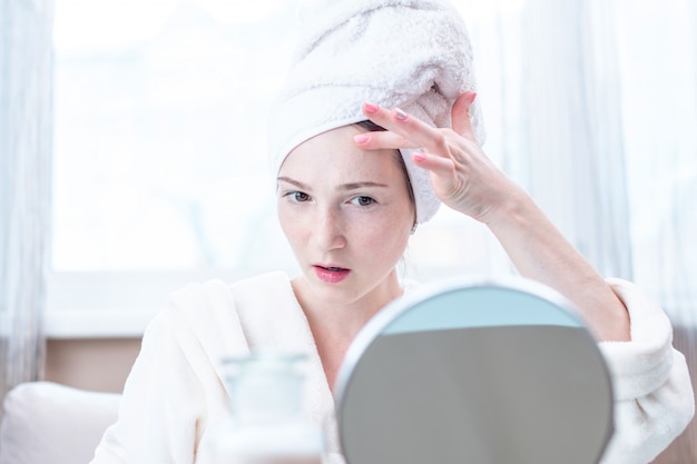 Beautiful happy woman with a towel on her head looking at her skin in a mirror. Hygiene and care for the skin