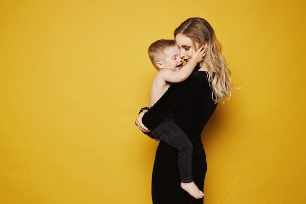 Beautiful and happy young blonde woman in fashionable black dress with cute little baby boy on her h...