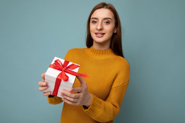 Beautiful happy young brunette woman isolated over colourful background wall wearing stylish casual clothes holding gift box and looking at camera