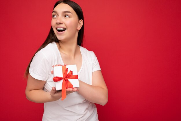 Beautiful happy young brunette woman isolated over colourful background wall wearing stylish casual clothes holding gift box and looking to the side