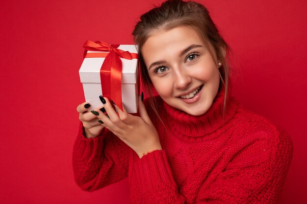 Beautiful happy young brunette woman isolated over colourful wall wall wearing stylish casual clothes holding gift box and .