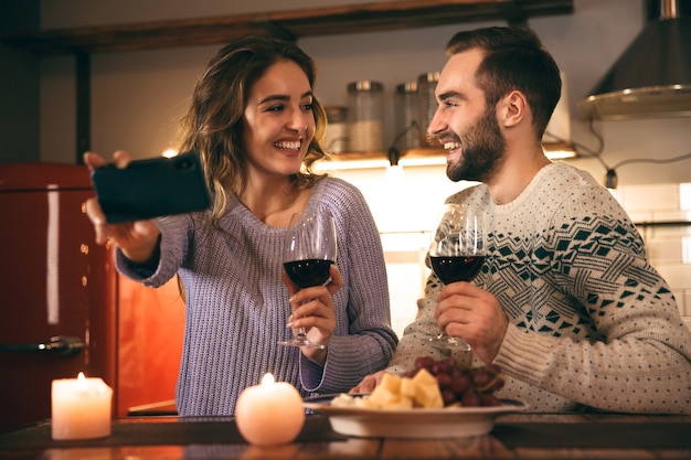 Beautiful happy young couple spending romantic evening together at home, drinking red wine, taking a selfie