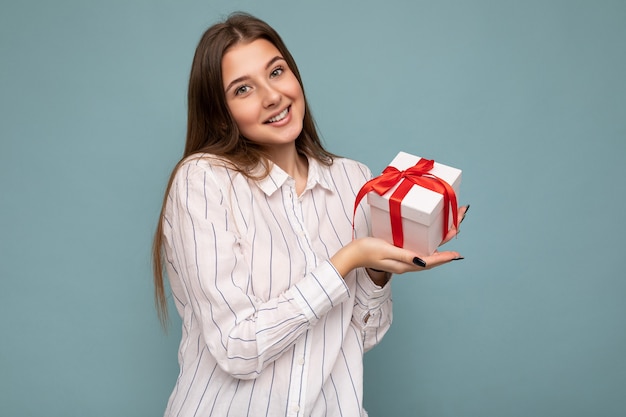 beautiful happy young dark blonde woman isolated over colourful background wall wearing stylish casual clothes holding gift box and looking at camera