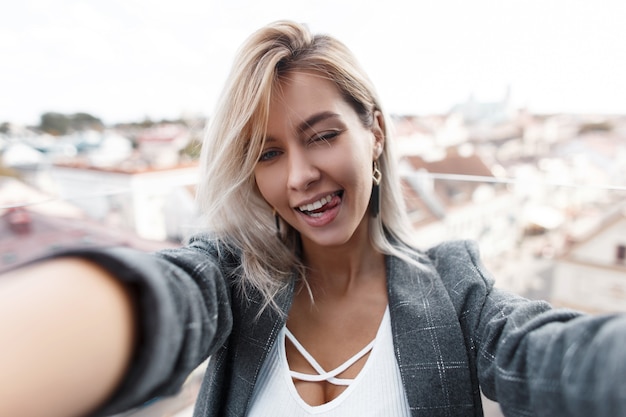 Photo beautiful happy young fashionable girl shows tongue and makes selfie in the city. woman is traveling and taking a photo on the phone.