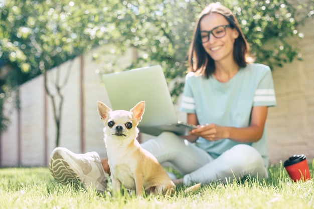 Beautiful happy young freelance woman with her little cute dog is working on a laptop outdoor.