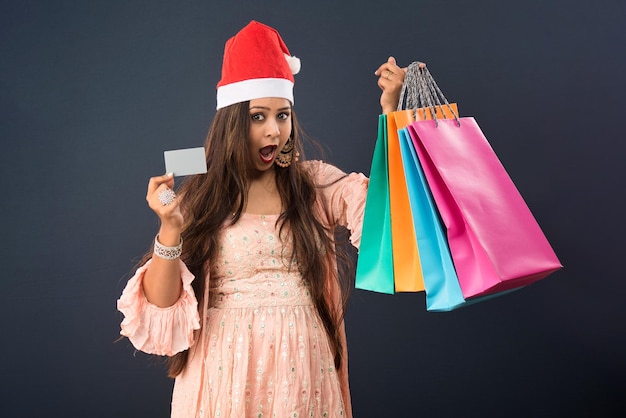 Beautiful Happy Young Girl in Santa hat holding gift bags and credit card on grey background Christmas shopping concept or buying presents for Christmas