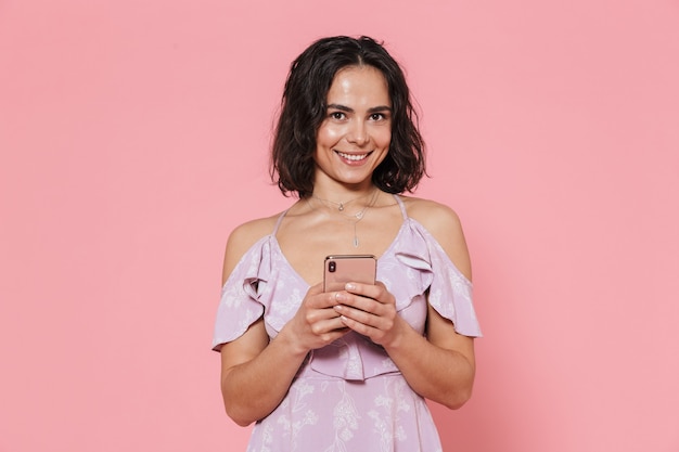 Bella ragazza felice che indossa un abito estivo in piedi isolato sul muro rosa, usando il telefono cellulare