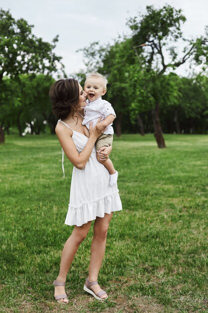 Beautiful and happy young mom in short white dress with a cute little stylish son on her hands posing at green park