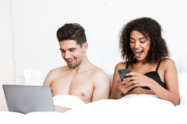 Beautiful happy young multiethnic couple relaxing in bed under blanket, man using laptop computer, woman holding mobile phone