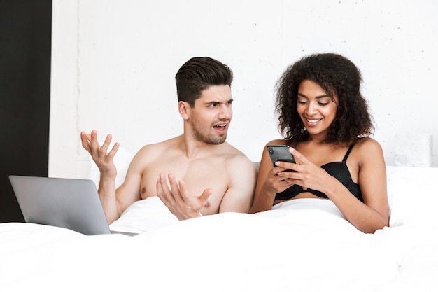 Beautiful happy young multiethnic couple relaxing in bed under blanket, man using laptop computer, woman holding mobile phone