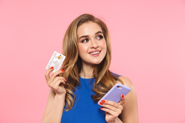 a beautiful happy young pretty woman posing isolated over pink wall holding credit card using mobile phone