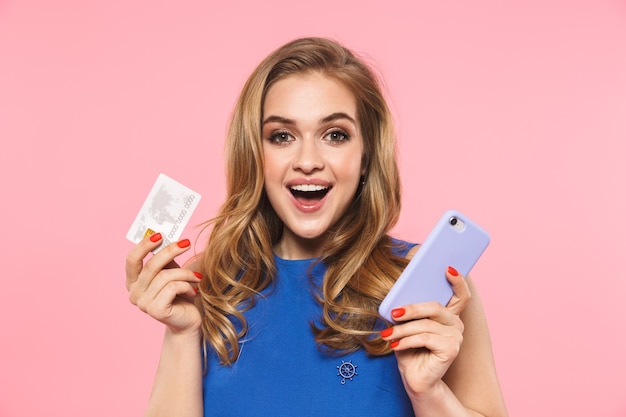 a beautiful happy young pretty woman posing isolated over pink wall holding credit card using mobile phone