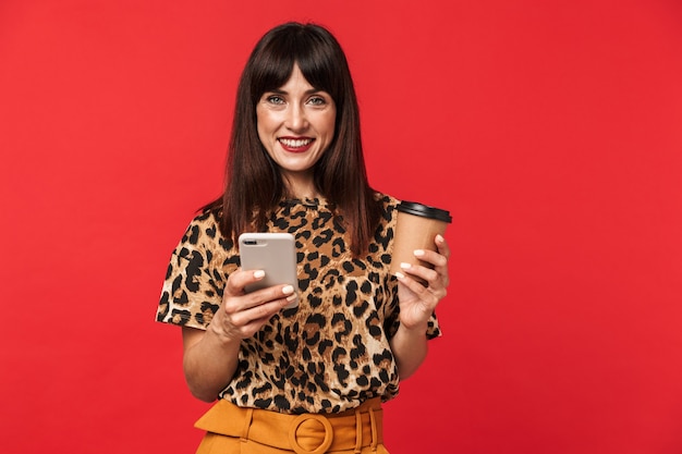 Bella giovane donna felice vestita con camicia stampata animale in posa isolata sul muro rosso che beve caffè utilizzando il telefono cellulare.