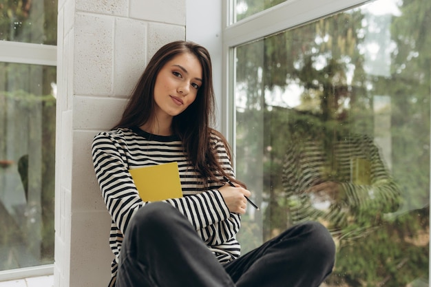 A beautiful happy young woman is sitting on the windowsill with a book in her hands