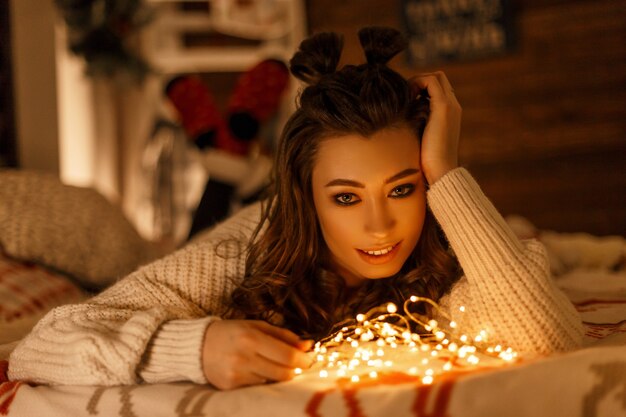 beautiful happy young woman in knitted sweater with festive lights on the bed on christmas eve