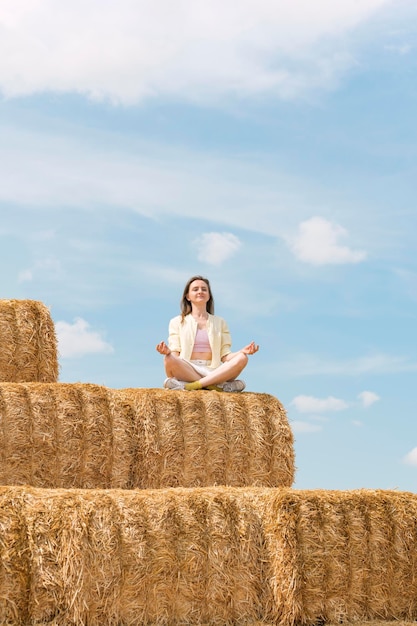 Beautiful happy young woman in lotus position on haystack and meditates Countryside holiday Blue sky background