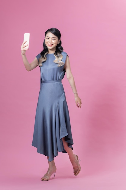 写真 ピンクの背景の上に立って、スマートフォンで自分撮りをしている美しい幸せな若い女性