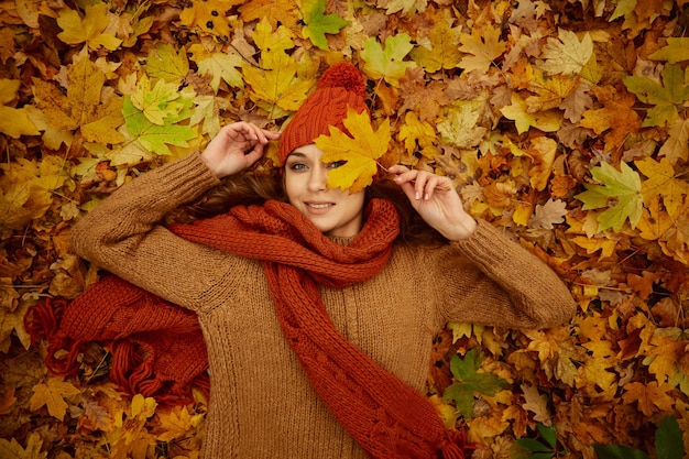 Bella giovane donna felice che indossa abiti lavorati a maglia sdraiati sulle foglie di acero autunnali. vista aerea dall'alto