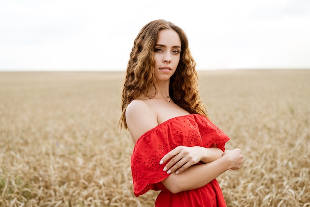 Beautiful happy young woman with curly hair in a red dress posing in a wheat field