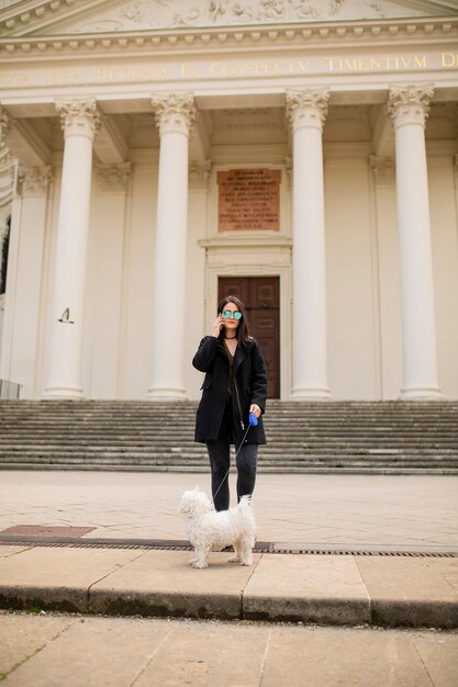 Beautiful happy young woman with cute small dog puppy using mobile phone on street of Vienna, austria