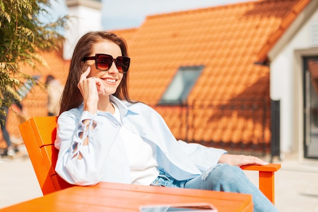 Beautiful happy young woman with a cute smile with vintage glasses in fashionable casual clothes with a shirt tshirt and jeans sits and rests outdoors on a sunny day