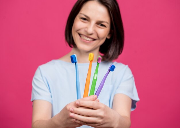 Beautiful happy young woman with different colored toothbrushes on blank pink background