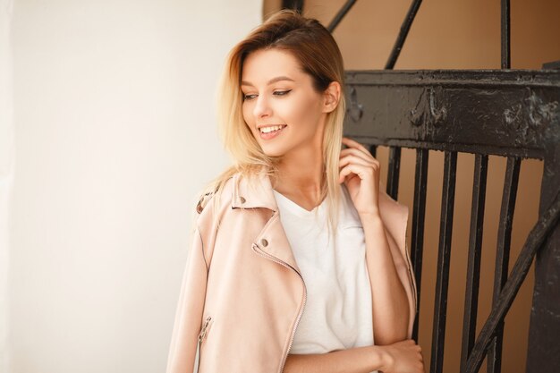 Красивая счастливая молодая женщина с улыбкой в стильной розовой куртке возле металлического забора на улице
