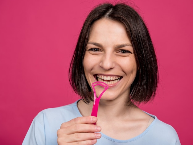 Beautiful happy young woman with tongue scraper on blank pink background