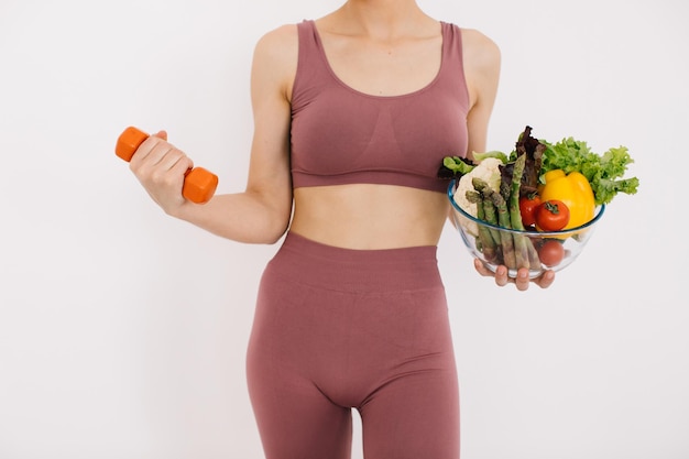 Beautiful happy young woman with tray of various healthy\
vegetables and showing her muscles with dumbbell isolated on the\
white background