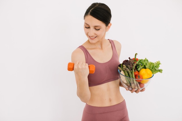 Beautiful happy young woman with tray of various healthy vegetables and showing her muscles with dumbbell isolated on the white background