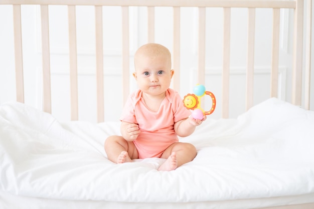 A beautiful healthy cheerful baby is sitting in a crib with\
white bed linen the child is holding a toy looking at the\
camera