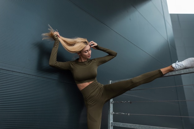 Красивая здоровая фитнес-блондинка со спортивным телом в зеленой тренировочной одежде с кроссовками делает упражнения на растяжку ног возле современного металлического здания в городе