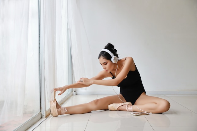 Beautiful healthy and sporty young asian woman in black sportswear sitting with headphone, listening music from mobile phone while training ballet dancing on white wall .technology and healthy concept