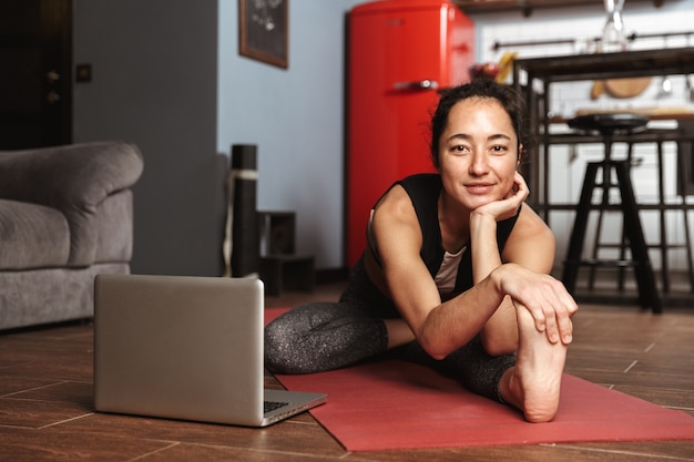 Beautiful healthy woman doing yoga exercises while sitting on a fitness mat at home, using laptop computer, stretching