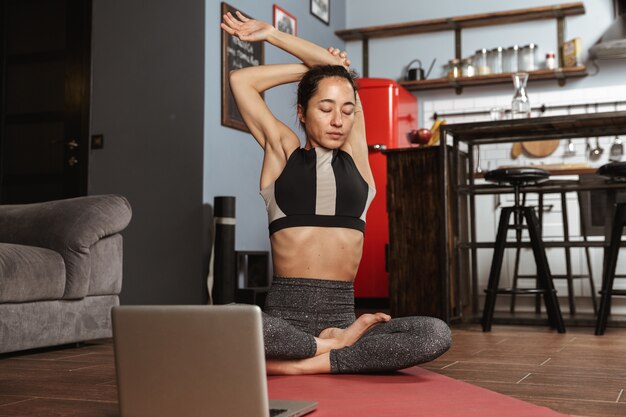 Фото Красивая здоровая женщина делает упражнения йоги, сидя на фитнес-коврике дома, используя портативный компьютер, растягивая