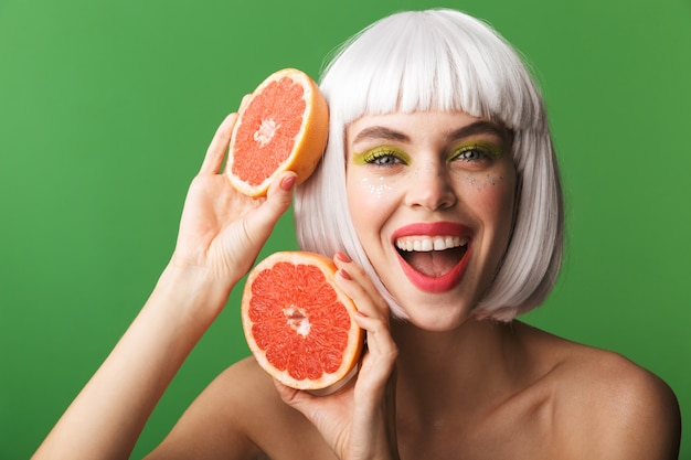Красивая здоровая молодая женщина топлес в коротких белых волосах стоит изолированно, показывая нарезанный грейпфрут
