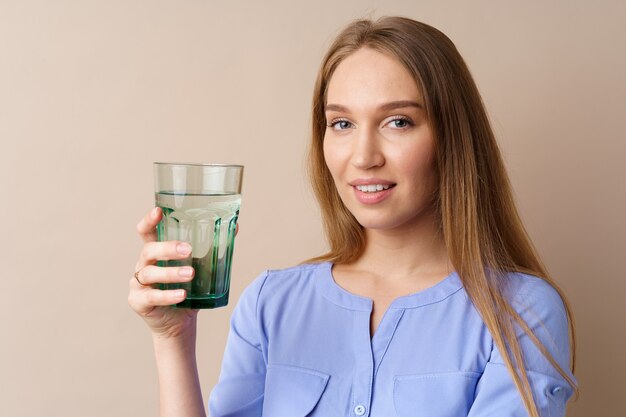 Beautiful healthy young woman drinking water from a glass on beige background