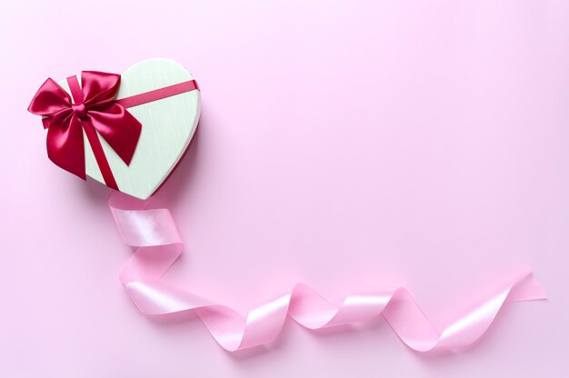 弓、コピースペースのあるピンクの背景にシルクのリボンが付いた美しいハート型のボックス。バレンタインデーへのギフト。