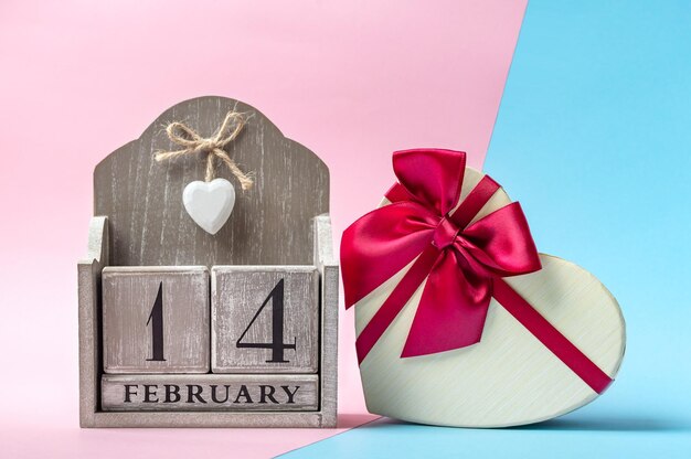 Beautiful heart shaped gift box with ribbon and wooden perpetual calendar on a modern two-tone background. Close-up. Concept of gift for your loved ones on Valentine's day on February 14