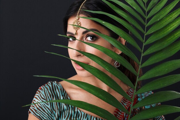 beautiful hindus girl with makeup wearing traditional indian saree dress and ethnic jewelry holding green plant isolated over black wall