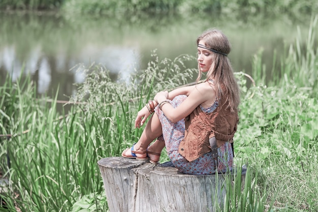 Beautiful hippie girl sitting on a stump near the pond
