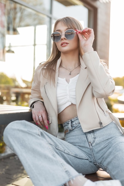 Beautiful hipster girl in fashion rock urban clothing with leather jacket top and jeans sitting on a bench and wearing cool fashion sunglasses on the street at sunset