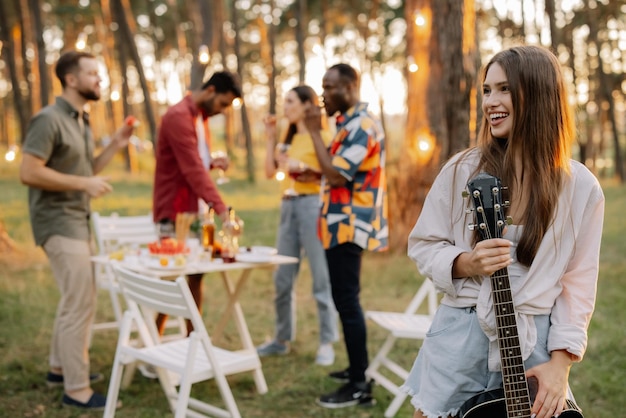 Photo beautiful hipster girl playing the guitar on the background of a dinner party outdoors