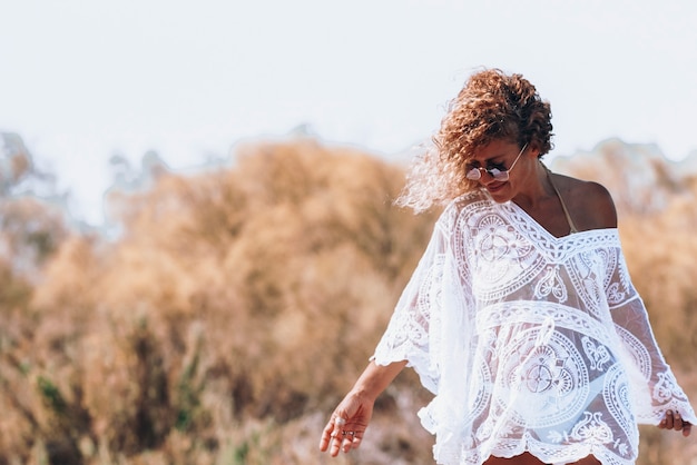 Beautiful hipster woman in curly hair, sunglasses and see\
through white dress looking down on hay field during summer tour.\
stylish woman in white dress enjoying summer vacations at\
countryside