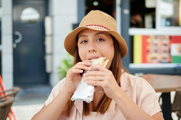 A beautiful Hispanic female in a hat eating a kebab in a cafe on a sunny day