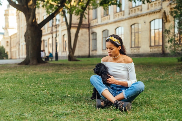 Beautiful hispanic woman in casual clothes sits outdoors on the lawn with a cute little dog