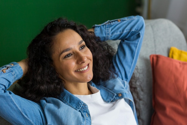 Beautiful hispanic woman daydreaming sitting on couch at home and looking out window happy woman