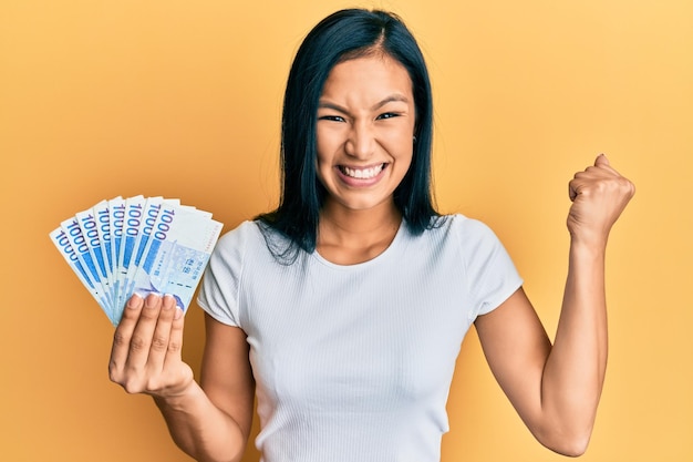 Beautiful hispanic woman holding 1000 south korean won banknotes screaming proud celebrating victory and success very excited with raised arm